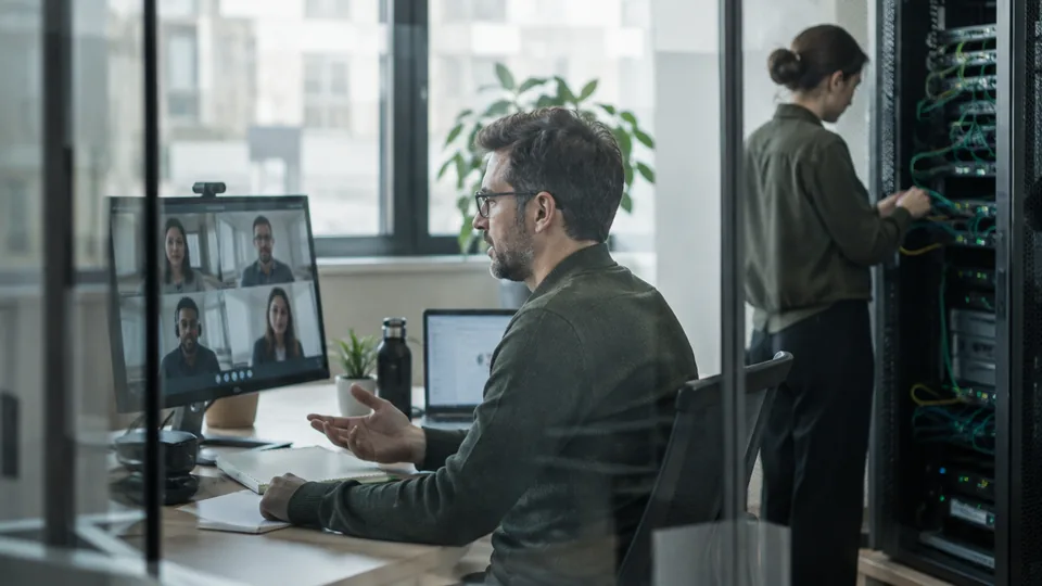 Distributed IT team coordinating branch security monitoring on a video call beside a network cabinet.